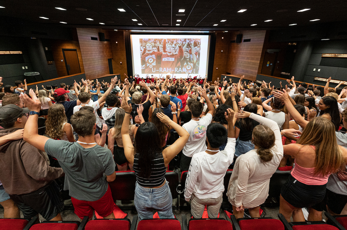 A large group of new UW-Madison students in Union South's Marquee Theatre sing and wave their arms in the air.