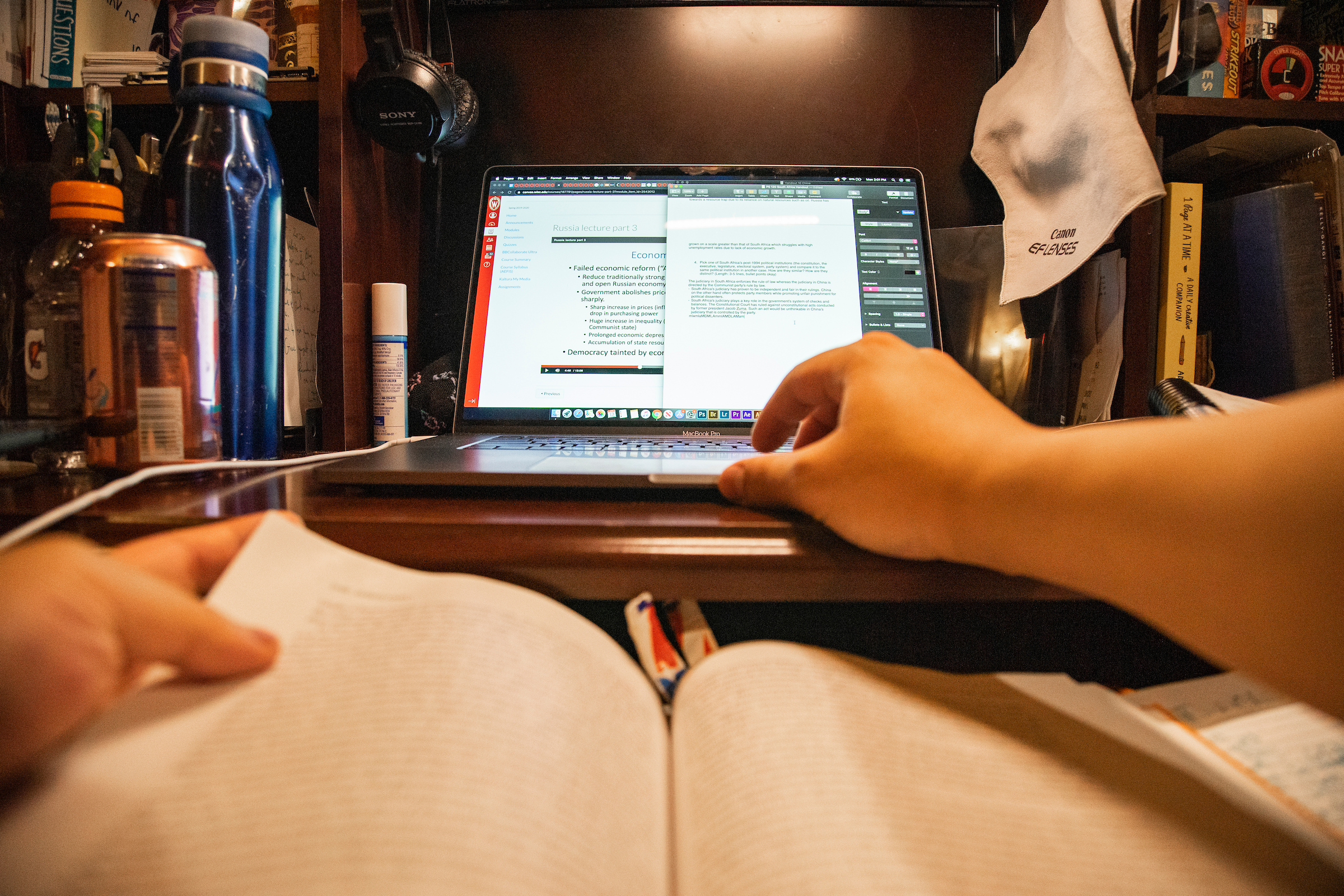 A student's point of view, with a book opened directly in the foreground and a laptop computer opened up on a desk in the background.